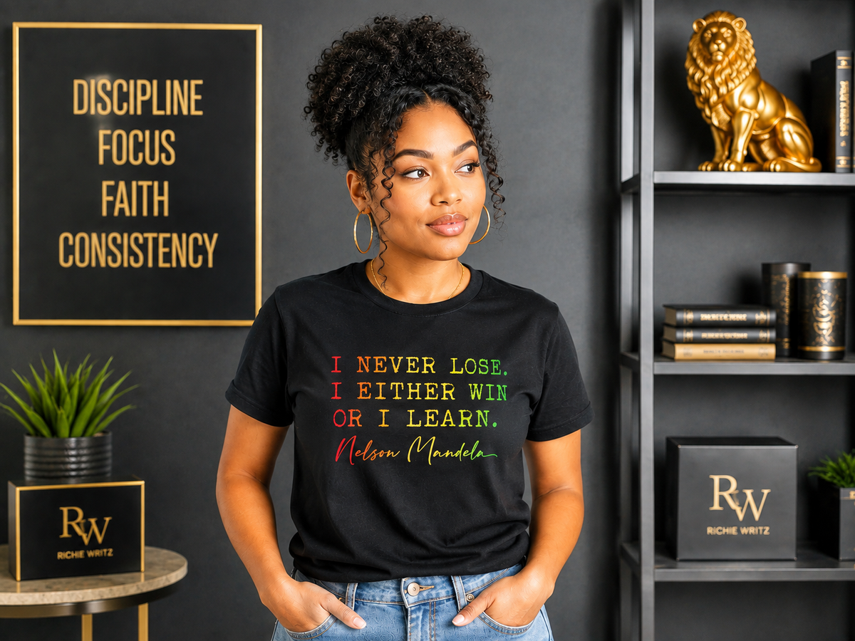 A woman wearing a black graphic t-shirt with a Nelson Mandela quote in red, yellow, and green text, standing in a styled room with a dark gray wall, gold-framed sign, and bookshelves.
