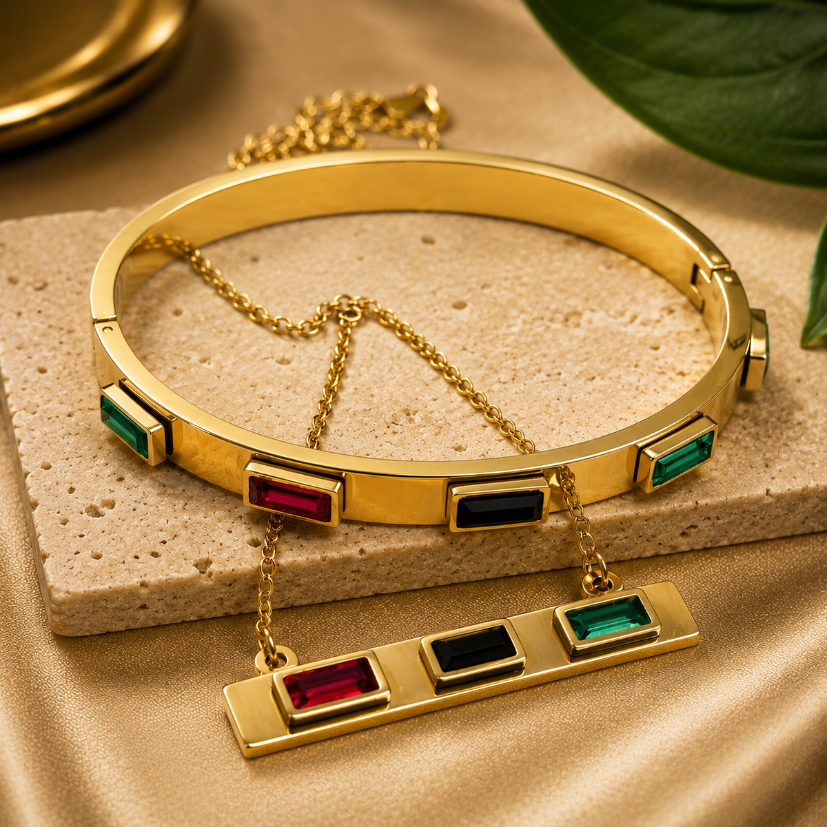 Gold-toned bangle necklace with rectangular multicolor gemstone accents and a matching drop bar pendant, displayed on a beige stone slab with satin fabric and a leaf in the background.